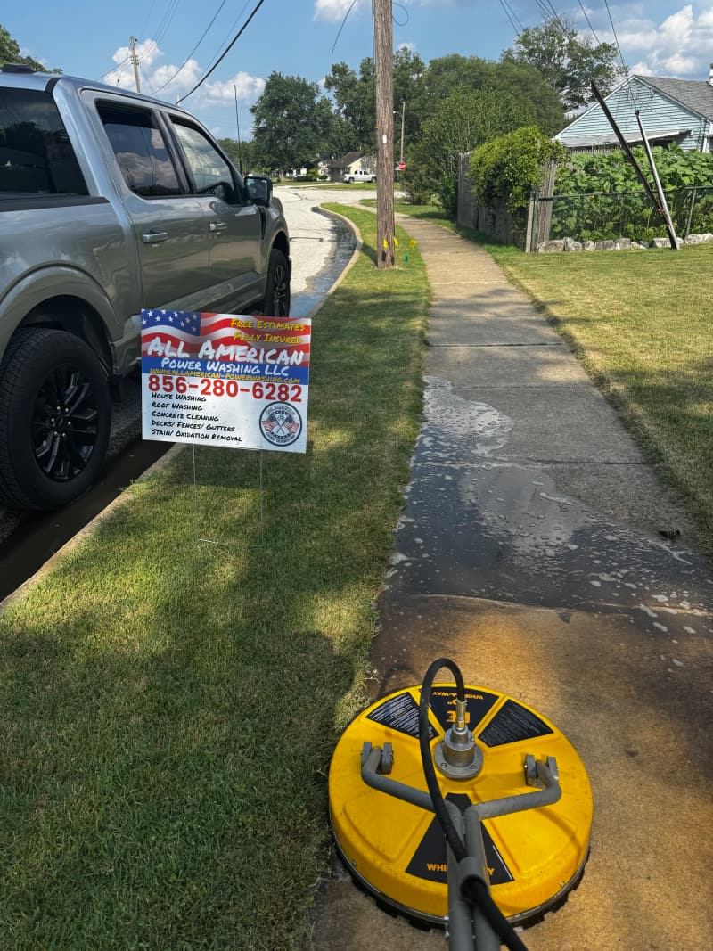 A yellow surface cleaner on a wet sidewalk beside an All American Power Washing yard sign